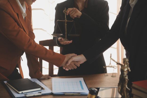 Advogados realizando handshake durante processo de due diligence jurídica em fusões e aquisições, com balança da justiça ao fundo.