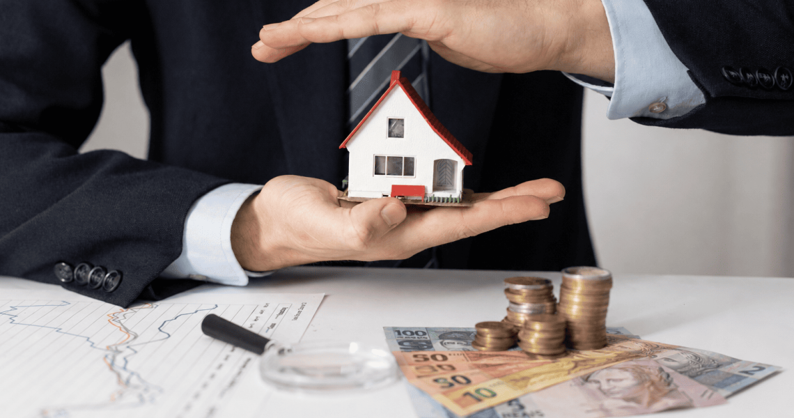 Empresário protegendo miniatura de casa com as mãos, com notas de reais, moedas e gráficos financeiros sobre a mesa, representando blindagem patrimonial.