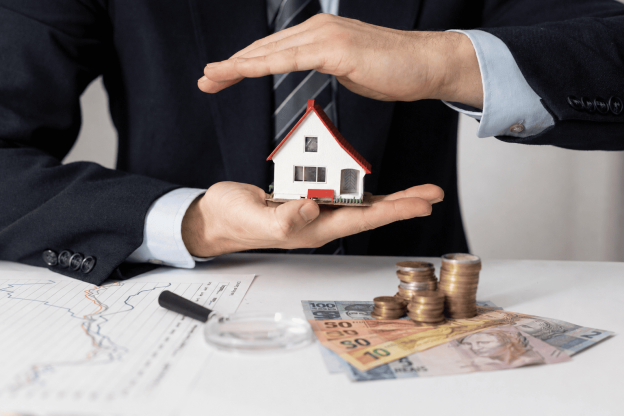 Empresário protegendo miniatura de casa com as mãos, com notas de reais, moedas e gráficos financeiros sobre a mesa, representando blindagem patrimonial.
