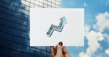 Mão segurando papel com seta de crescimento em frente a prédio empresarial, representando crescimento empresarial jurídico.